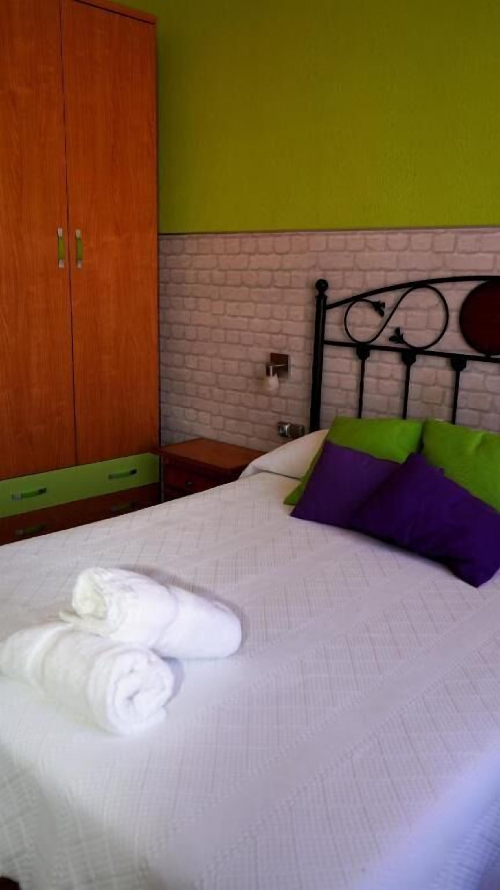Una cama bien hecha con ropa de cama blanca y toallas enrolladas, un armario de madera y una pared con diseño de ladrillo.