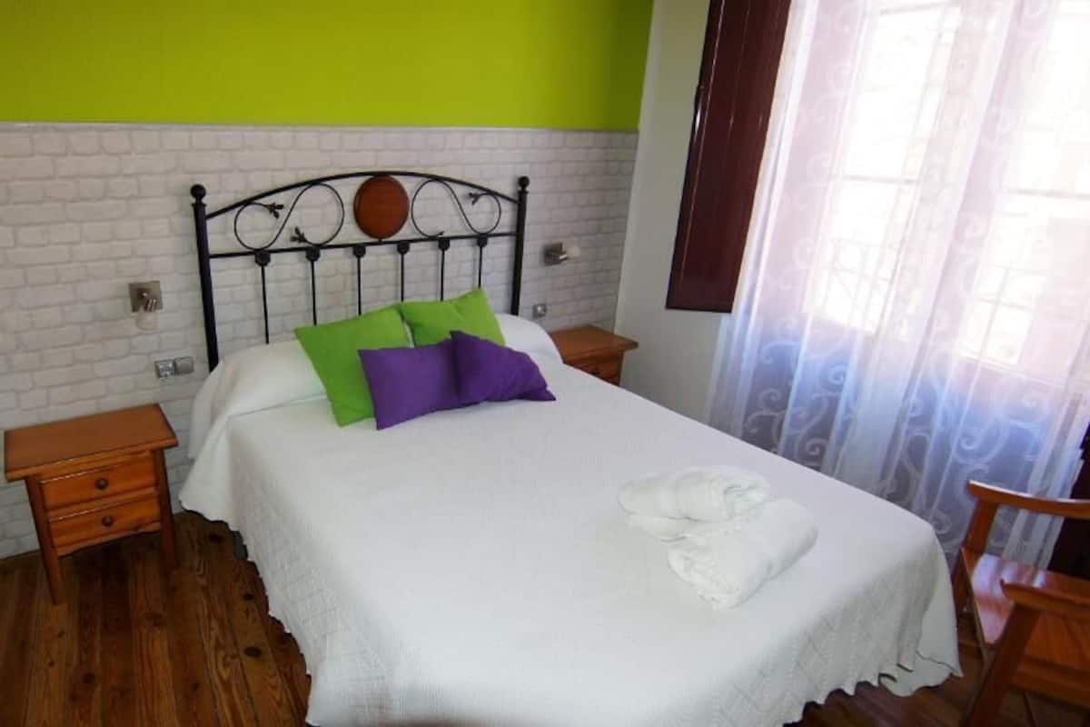 Un letto rifatto con una coperta bianca e cuscini verdi e viola, un comodino in legno e una sedia di legno.