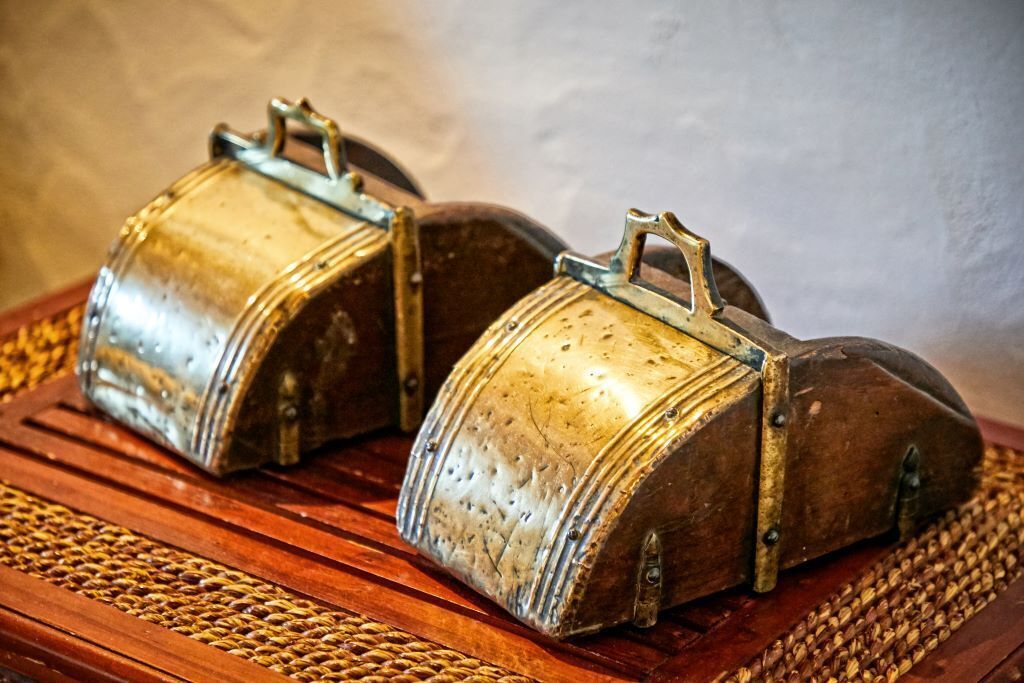 Two vintage metal objects with handles, possibly antique containers, placed on a wooden surface with a woven mat underneath.
