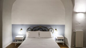 Una camera da letto con un ampio soffitto a volta, un letto con lenzuola bianche, due comodini con lampade e un termosifone sulla parete.