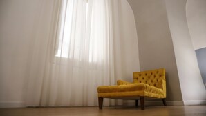 Una stanza con una poltrona gialla, un'ampia finestra con tende leggere e un soffitto alto ad arco.