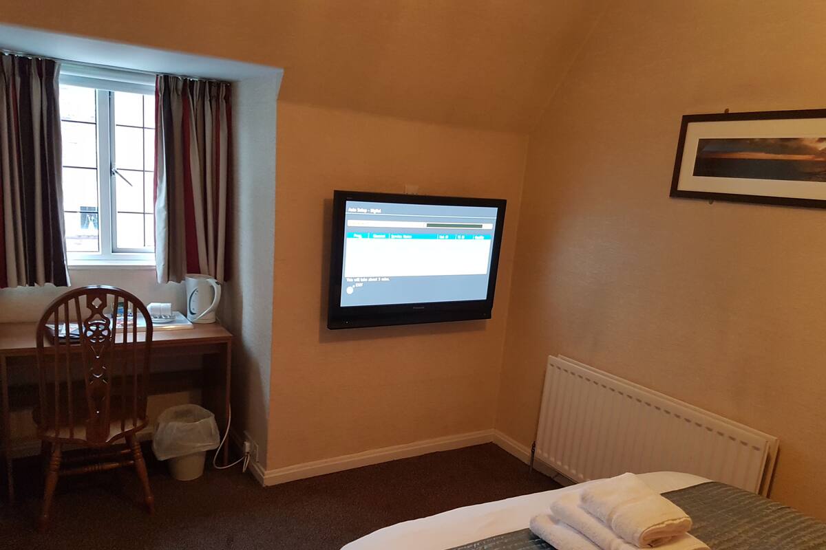 A hotel room with a bed, a desk with a computer, a chair, a radiator, and a window with curtains.