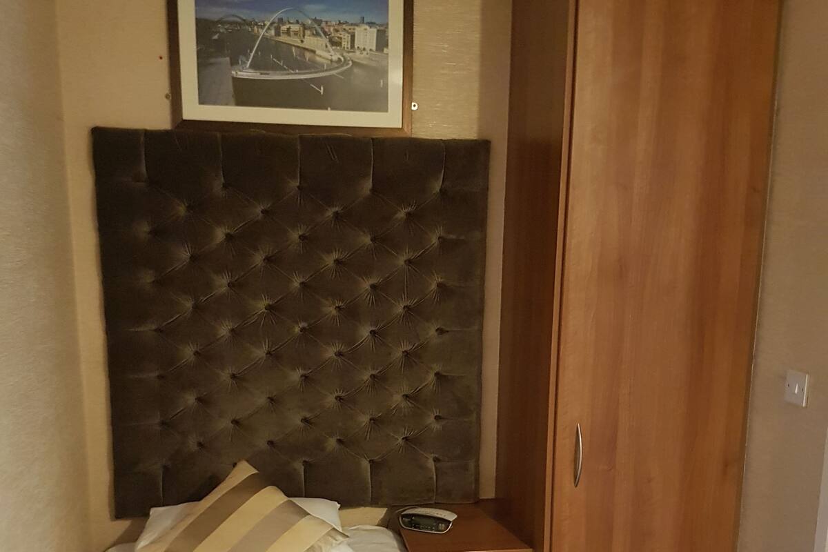 A neatly made bed with a striped pillow, a bedside table with a radio, a framed picture above the bed, and a wooden wardrobe.