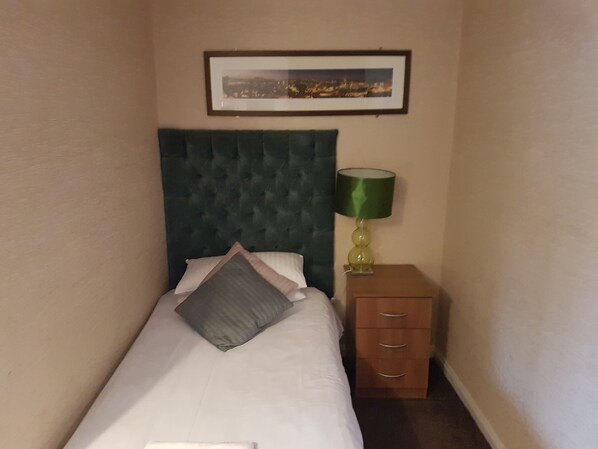 Una cama pequeña y ordenada con cabecera verde y una sola almohada. Una lámpara verde está sobre una mesita de noche de madera junto a la cama.