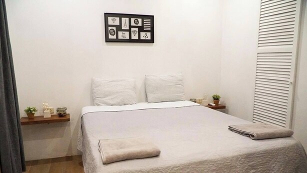 A neatly made bed with a white duvet, a small bedside table with a plant, and a wall-mounted picture frame. - Condominios Las Navieras (Tamarindo)