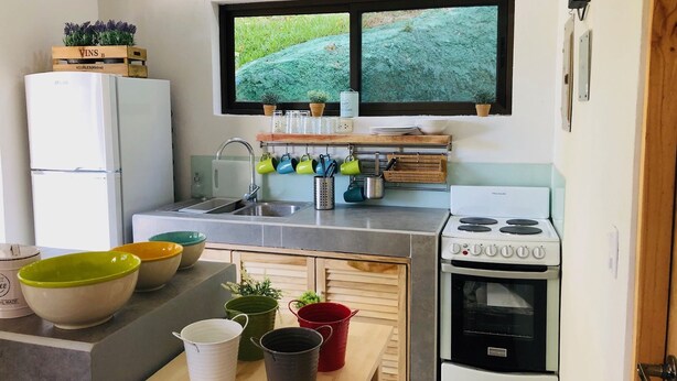 A compact kitchen with a refrigerator, sink, stove, and window overlooking greenery. - Condominios Las Navieras (Tamarindo)