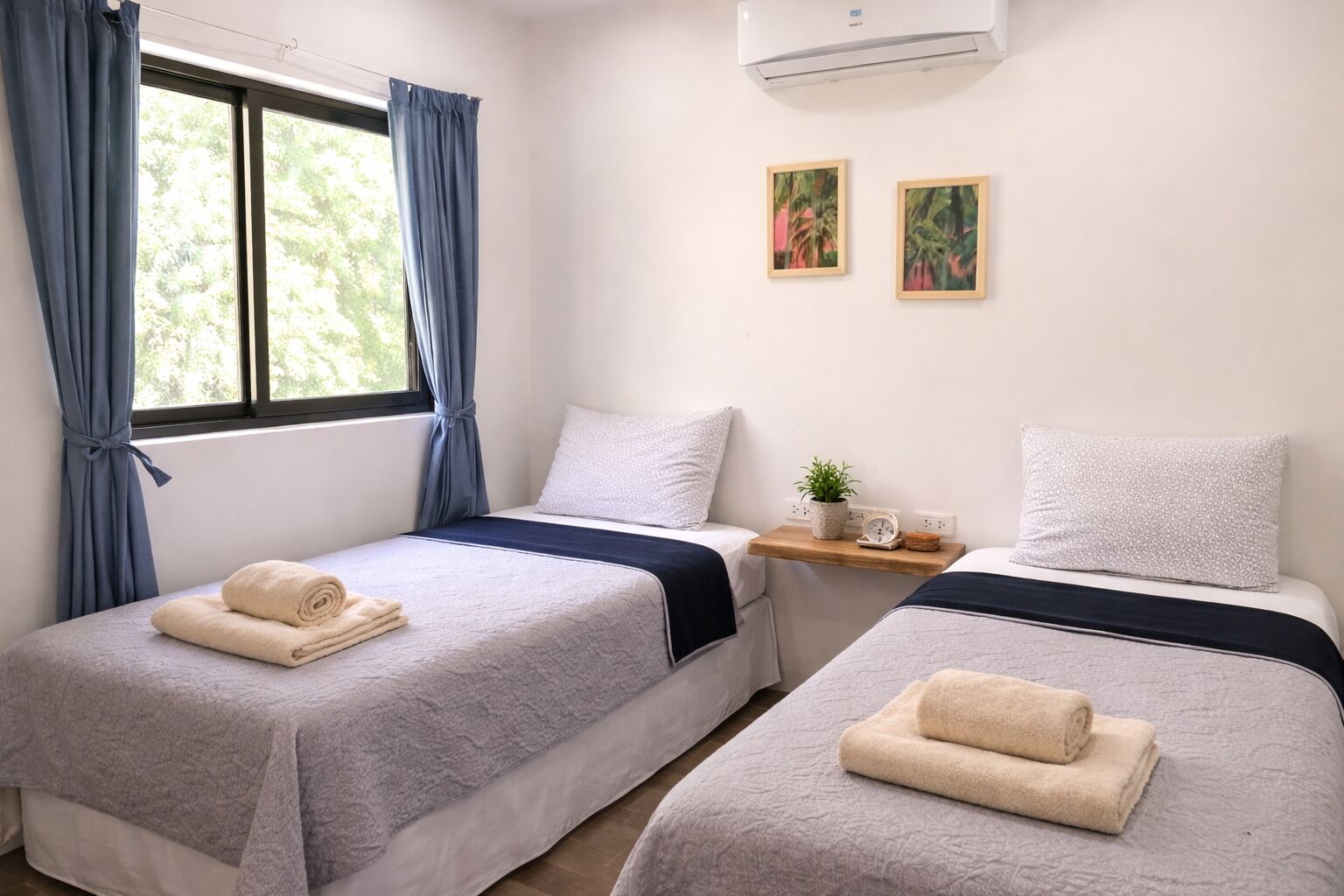 A hotel room with two beds, each with a rolled towel, a small table with a plant and a clock, and a window with curtains.