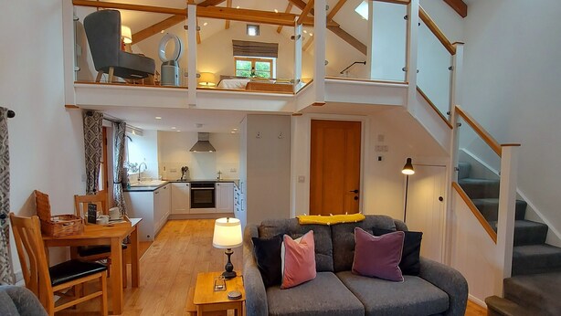 A spacious loft with a high ceiling, exposed wooden beams, a dining area, a sofa set, and a staircase leading to an upper level. - Spacious, luxury,  romantic Barn for couples near Exmoor & Coast  (Bratton Fleming, Barnstaple)