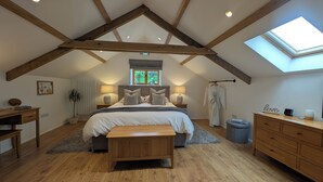 A bedroom with a sloped ceiling, wooden beams, a skylight, a bed with pillows, bedside lamps, a bench, a dresser, and a radiator. - Spacious, luxury,  romantic Barn for couples near Exmoor & Coast  (Bratton Fleming, Barnstaple)