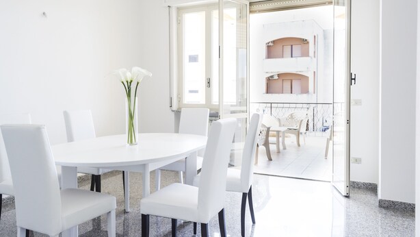 Zona pranzo moderna con tavolo e sedie bianche, un vaso con fiori e vista su un edificio residenziale attraverso una porta in vetro.