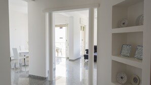 Un corridoio ampio e luminoso con pavimento lucido, che conduce a una sala da pranzo con sedie bianche e un tavolo. Si notano delle mensole con piatti decorativi a destra.