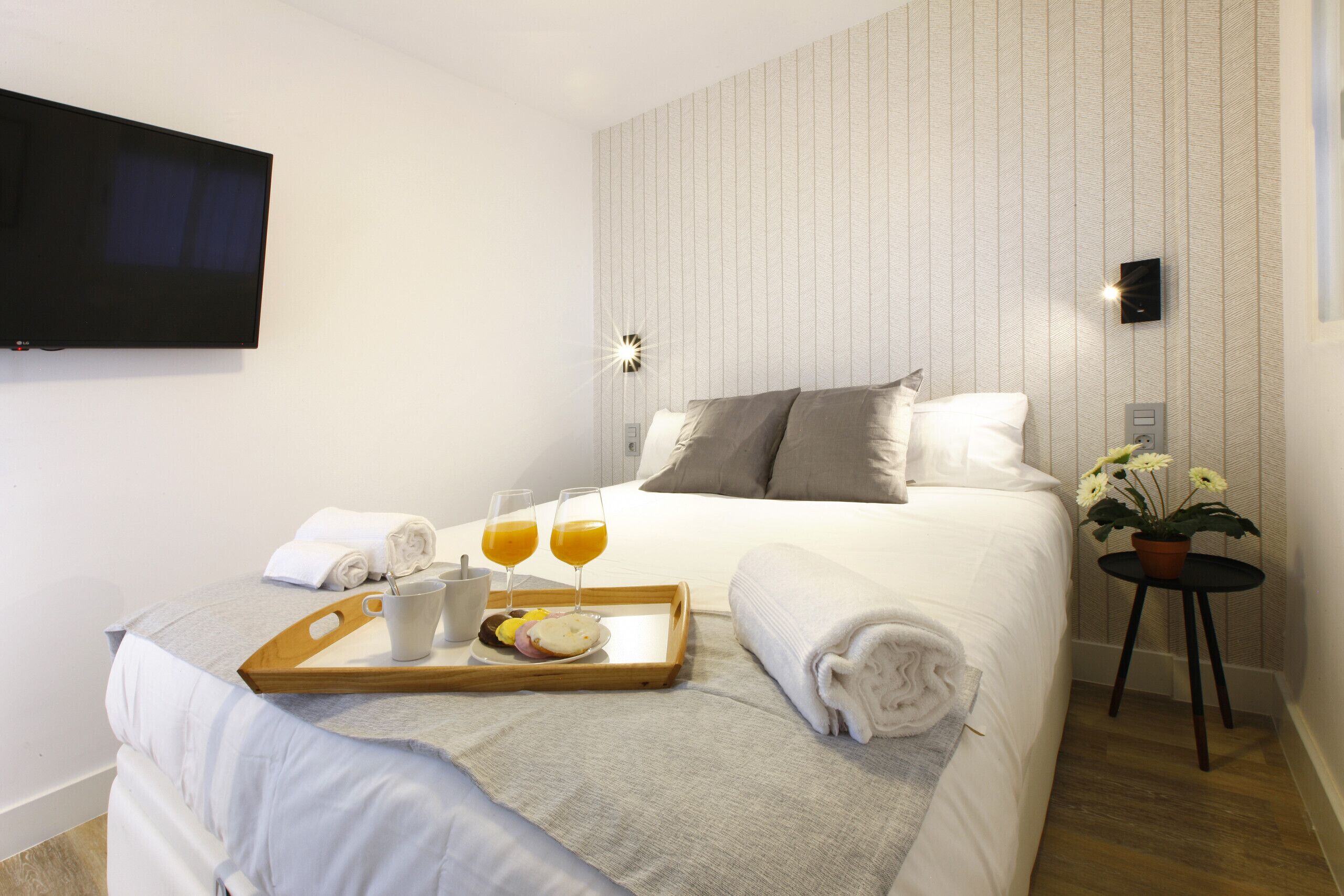 A neatly made bed with pillows, a tray with breakfast items, a bedside table with a plant, and a wall-mounted TV.