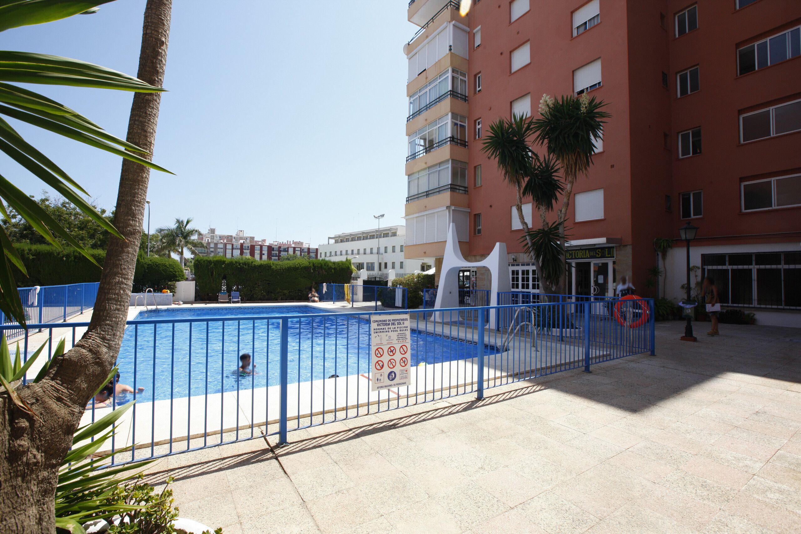 A swimming pool with a blue fence, a sign with safety instructions, and a building with balconies in the background.