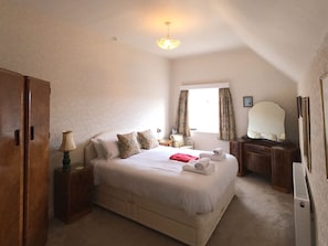 Un dormitorio con cama, mesitas de noche, armario, ventana con cortinas, espejo y candelabro.