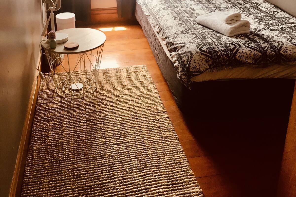 Una habitación pequeña y luminosa con una cama individual, una mesita de noche, una pequeña mesa redonda con un florero y un lavamanos en la pared.