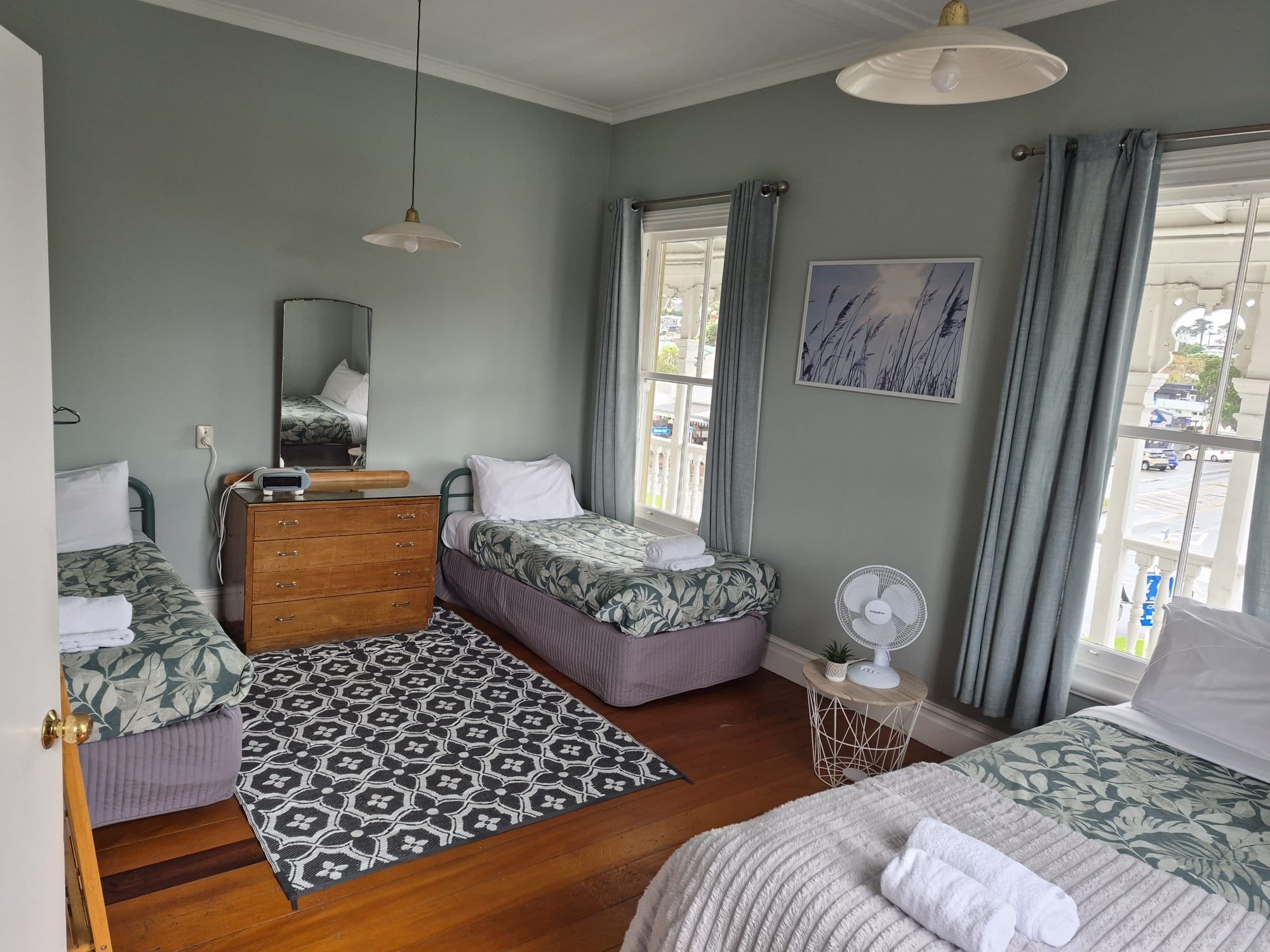 Une chambre avec deux lits, une commode, un miroir, un ventilateur et une fenêtre avec des rideaux.