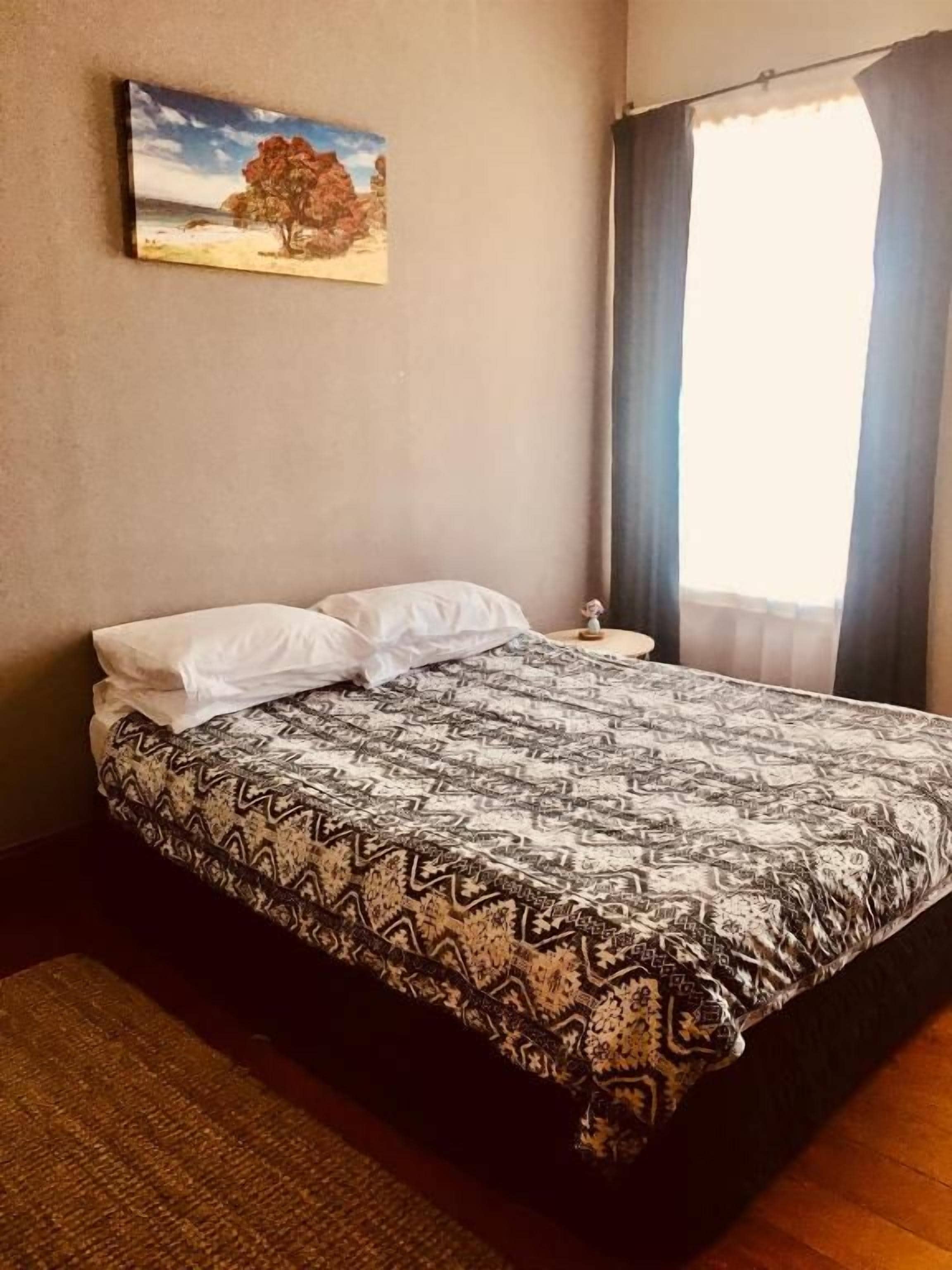 Un dormitorio con una cama, una mesita de noche, un cuadro en la pared y una ventana con cortinas.
