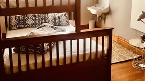 Una cama litera con almohadas estampadas, una mesita de noche, un lavabo blanco y un espejo en una habitación con ventana y lámpara colgante.