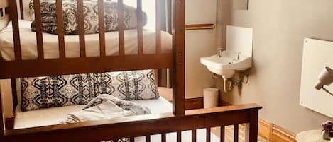 Un lit superposé avec des oreillers à motifs, une table de chevet, un lavabo blanc et un miroir dans une pièce dotée d’une fenêtre et d’un luminaire suspendu.