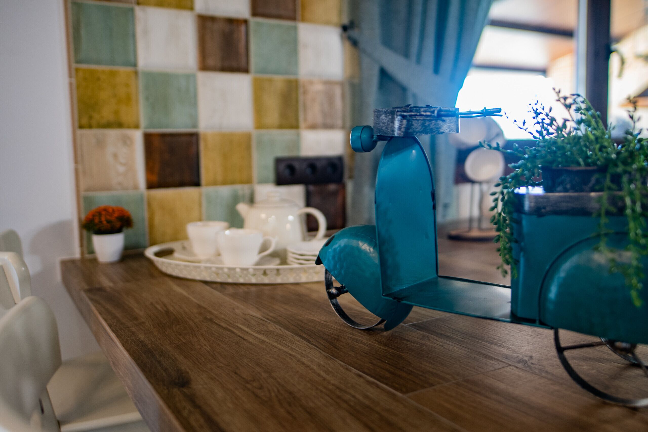 A teal espresso machine on a wooden table with a checkered tile backsplash and a potted plant.