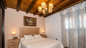 Un dormitorio con cabecera de madera, cama con cubrecama blanco texturizado, dos mesitas de noche con lámparas, un cuadro floral sobre la cama y una ventana con cortinas florales.