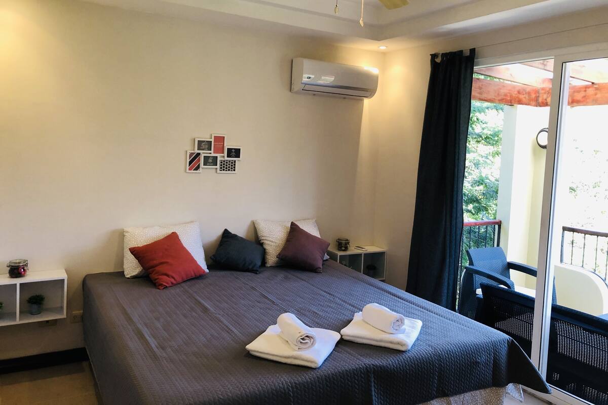 A hotel room with a bed, a ceiling fan, an air conditioning unit, a small table with a lamp, and a view of the outdoors through a sliding door.