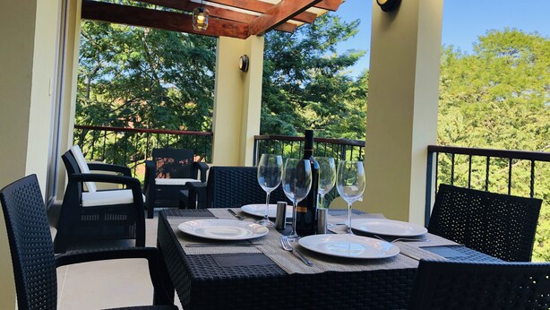 Uma área de refeições ao ar livre com pérgola, móveis de vime e uma mesa posta para quatro, com taças de vinho e pratos.