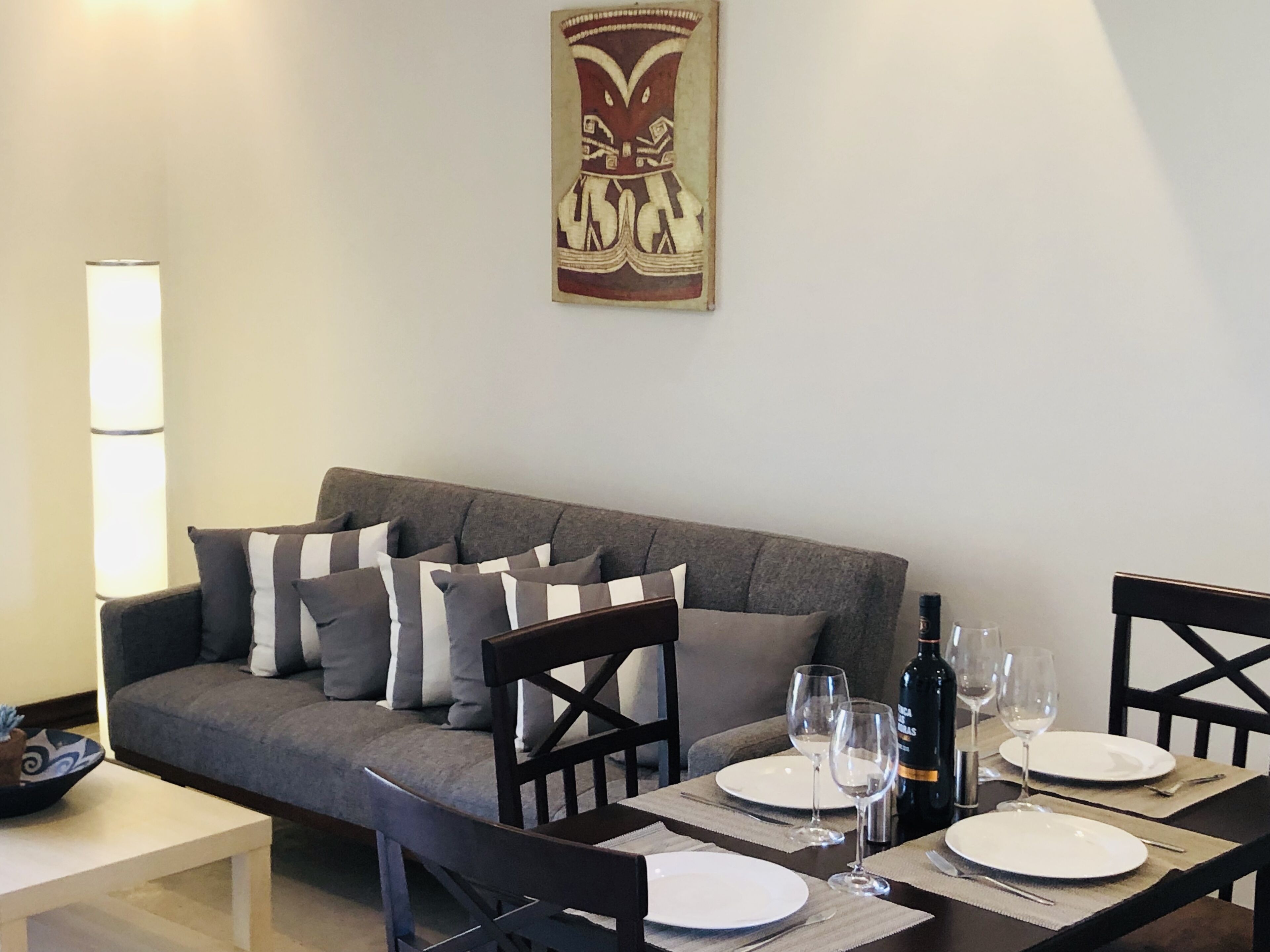 Un área de comedor con un sofá gris, almohadones a rayas blancas y negras, una mesa de madera con platos blancos y copas de vino, y una botella de vino.