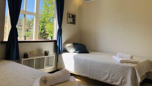 Quarto de hotel com duas camas, janela com cortinas, ar condicionado, um pequeno armário com objetos e vista para o verde lá fora.