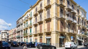 Um edifício de vários andares com varandas, um letreiro com 'TOMASINA' e carros estacionados na rua.