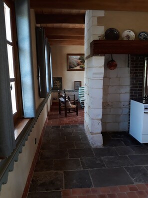 Un pasillo estrecho con suelo de piedra, una puerta de madera y una ventana a la izquierda. Se ve una silla y una mesa a lo lejos, y una chimenea de ladrillo con platos decorativos encima.