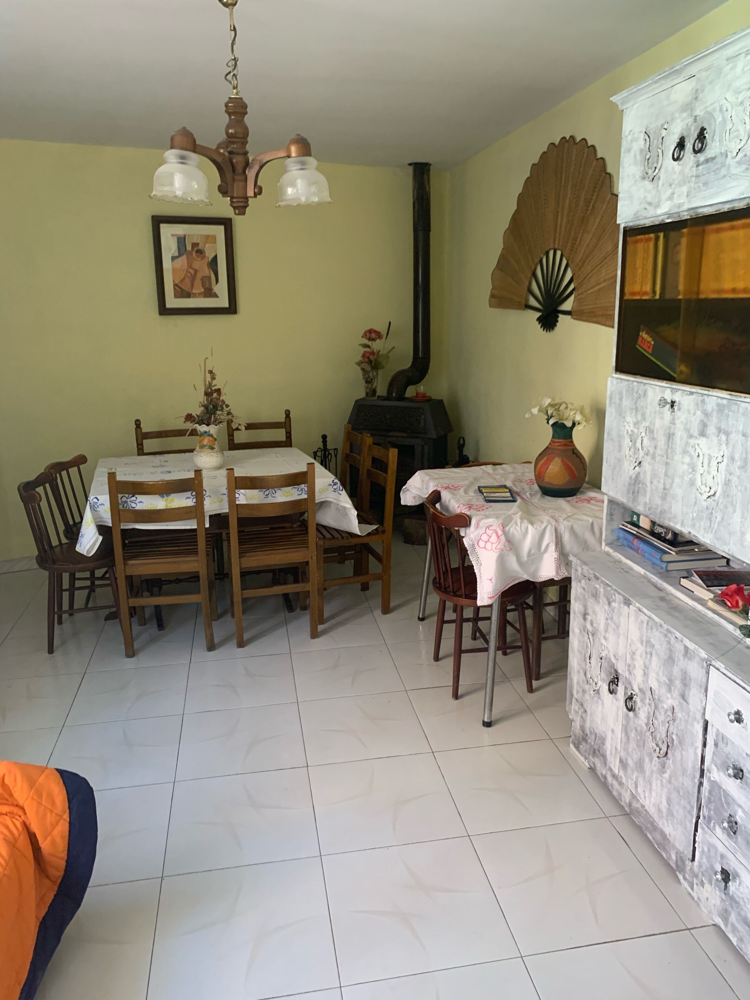 Un coin repas comprenant une table et des chaises en bois, un lustre, un poêle et un meuble contenant des livres et des objets décoratifs.