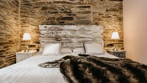Une chambre à coucher au style rustique, avec un mur en pierre, une tête de lit en bois, deux lampes de chevet et un plaid en fourrure sur le lit.