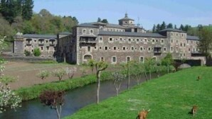 Un grand bâtiment en pierre, doté de nombreuses fenêtres et d’une tour centrale, entouré d’une zone herbeuse et bordé par une rivière.
