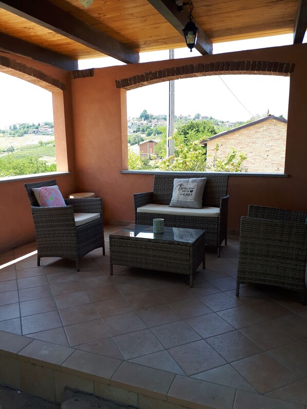 Ein überdachter Patio-Bereich mit Korbmöbeln, einem Glastisch und Blick auf einen Weinberg und Häuser.