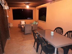 Eine Terrasse im Freien mit Esstisch, Stühlen, Korbmöbeln und Blick auf den Nachthimmel durch ein Fenster.