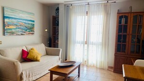 Una sala de estar con un sofá color beige, una mesa de centro, una estantería y un ventanal con cortinas translúcidas.