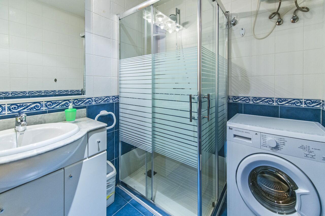 A bathroom with a glass shower enclosure, a washing machine, and a sink with a marble countertop.