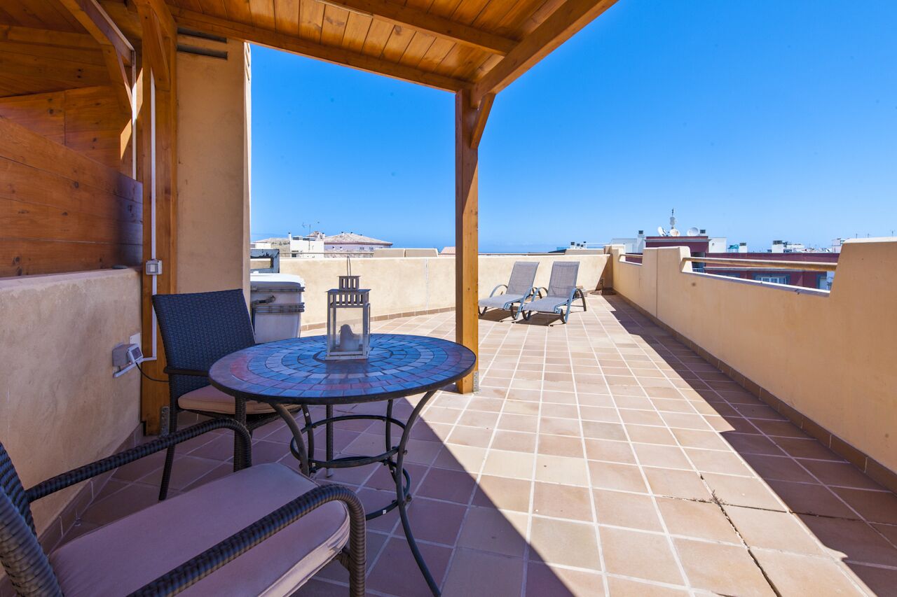 A covered outdoor seating area with a round table and chairs, a view of the sea, and a tiled floor.