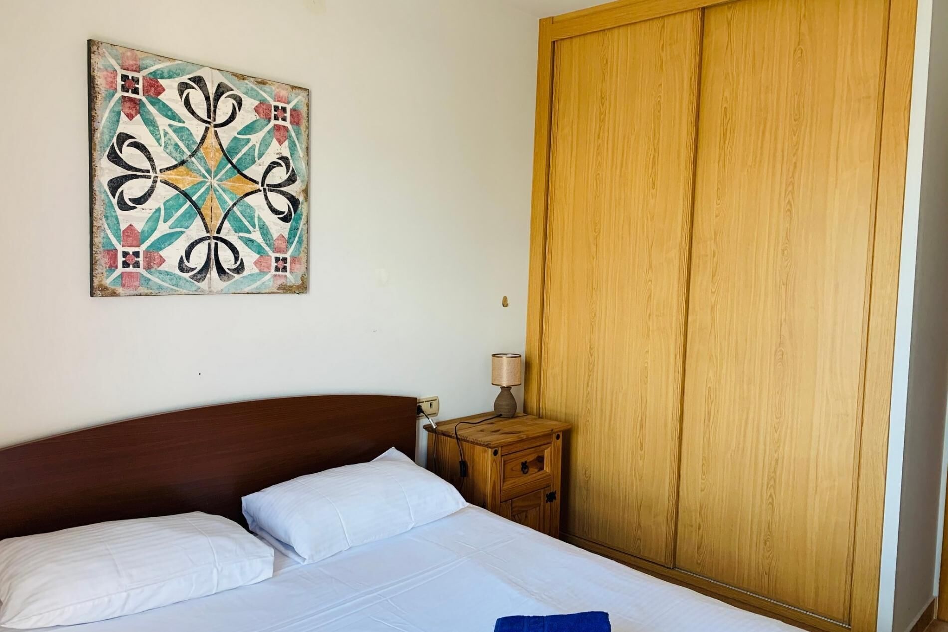 A bedroom with a bed, a wooden wardrobe, a nightstand, a lamp, and a wall art piece.
