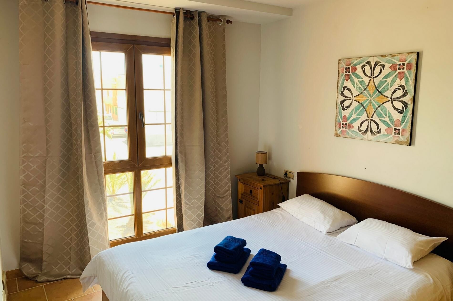 Habitación de hotel con cama, dos almohadas, mesita de noche con lámpara y ventana con cortinas.