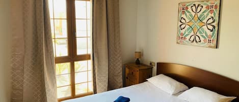 Habitación de hotel con cama, dos almohadas, mesita de noche con lámpara y ventana con cortinas.