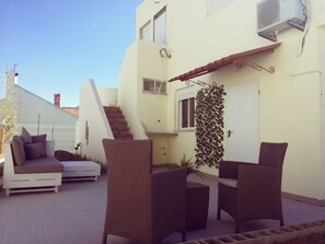 Un espacio al aire libre con sillas de mimbre y un sofá blanco, una escalera y un edificio blanco con unidades de aire acondicionado.