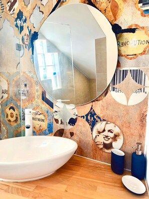 Un baño con un espejo redondo, un lavamanos blanco y suelo de madera. La pared presenta un diseño de mosaico con diversos patrones y texto.