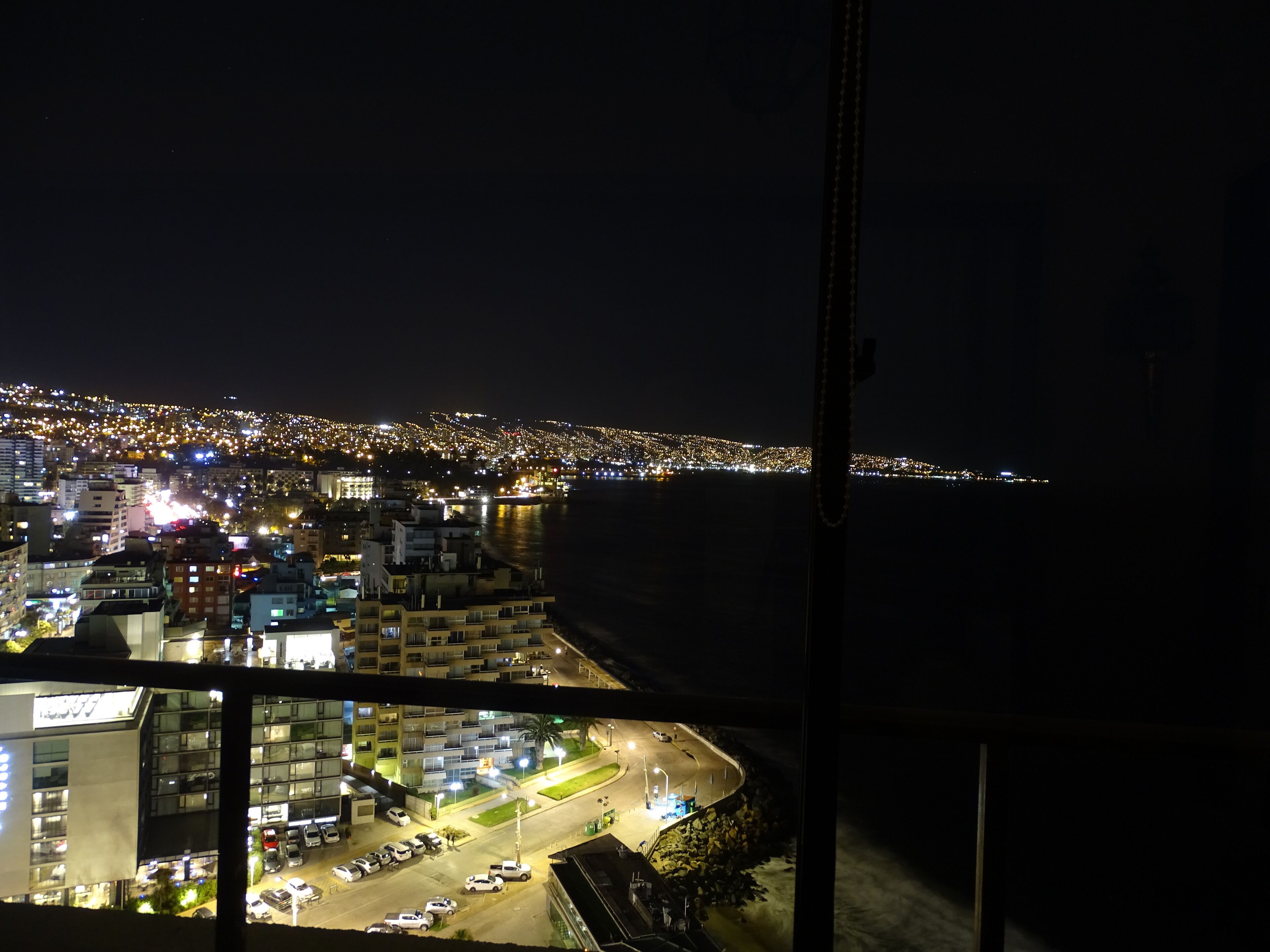 Une vue nocturne d’une ville, avec en arrière-plan une zone côtière, des bâtiments illuminés et un parking.