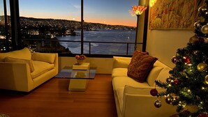Een moderne woonkamer met een groot raam dat uitkijkt over een stadsgezicht en water bij zonsondergang.