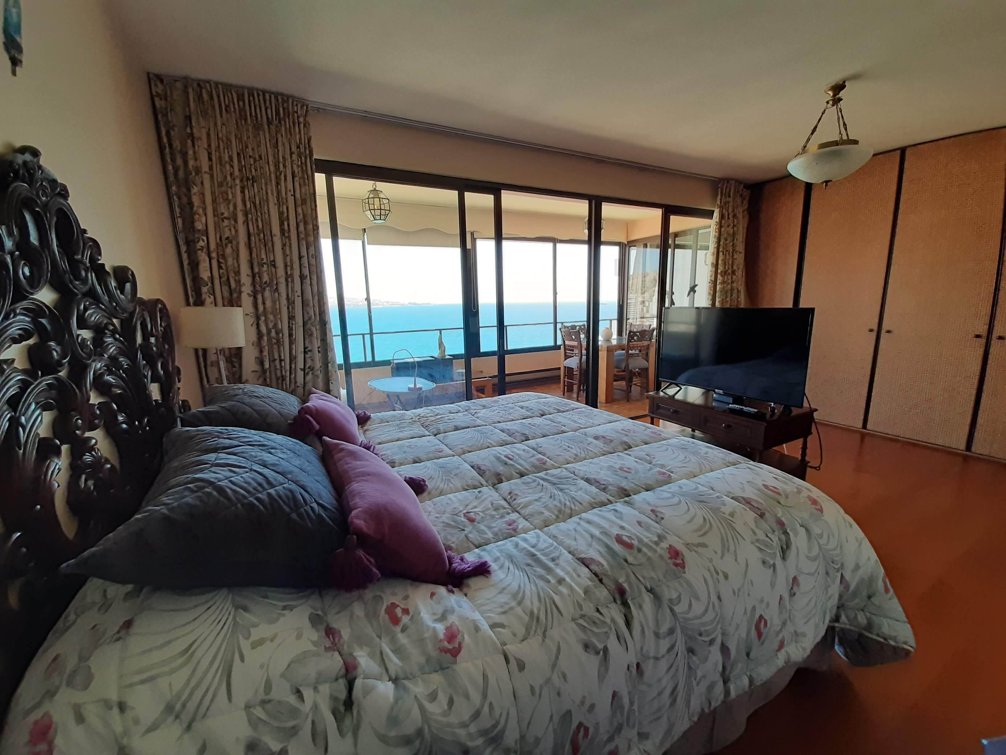 A bedroom with a large bed, a TV, and a balcony with a view of the sea.