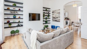 A modern living room with a grey sofa, a glass coffee table, and shelves with decorative items. - Luxury 4bed home 2min from High Street Ken/Olympia (London)