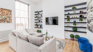 A modern living room with a grey sofa, a glass coffee table, and a blue chair. There are shelves with decorative items and a flat-screen TV mounted on the wall. - Luxury 4bed home 2min from High Street Ken/Olympia (London)
