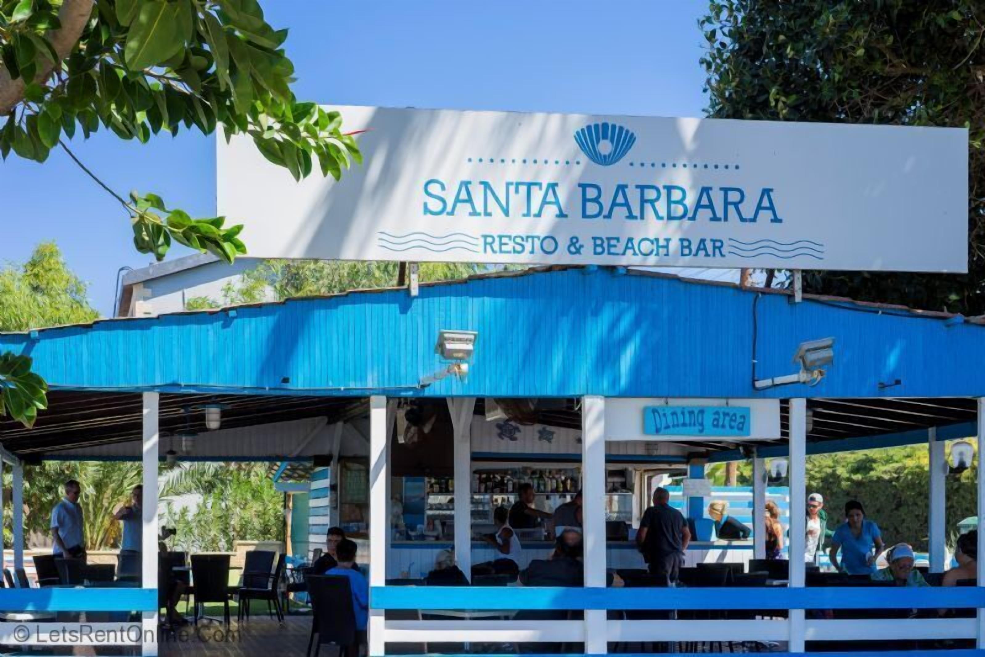 Et overdækket udendørs spiseområde ved Santa Barbara Resto & Beach Bar med et blåt tag og hvide søjler.